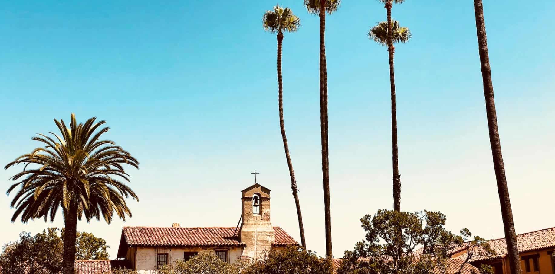 A picture of a Catholic California mission surrounded by palm trees and a blue sky backdrop.